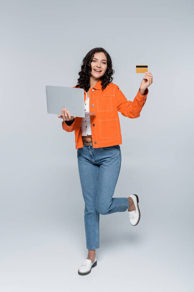 полная длина счастливой женщины в оранжевой куртке держа ноутбук и кредитную карту на серый 