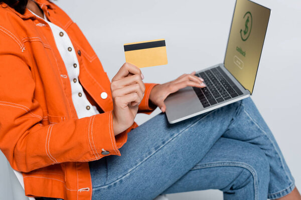 обрезанный вид женщины в оранжевой куртке с помощью ноутбука с кэшбэком буквы и проведение кредитная карта изолированы на серый 