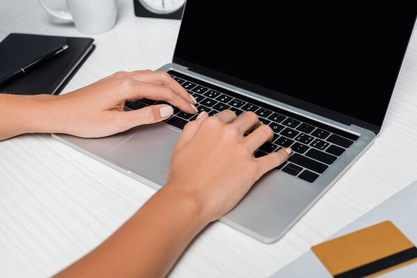 cropped view of woman typing on laptop with blank screen near credit card on white desk