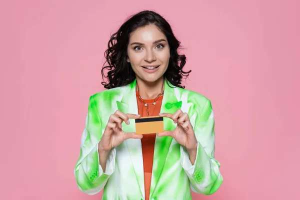cheerful young woman in tie-dye blazer holding credit card isolated on pink