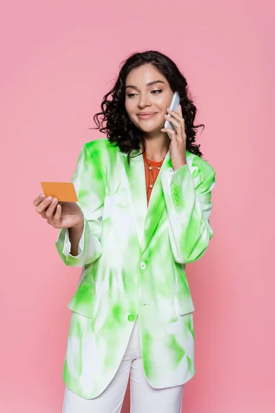 smiling young woman in tie dye blazer holding credit card and talking on smartphone isolated on pink