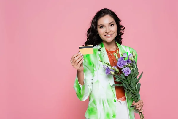 smiling young woman in tie dye blazer holding credit card and flowers isolated on pink