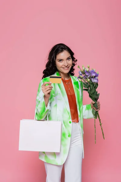 smiling young woman in tie dye blazer holding credit card, shopping bag and flowers isolated on pink