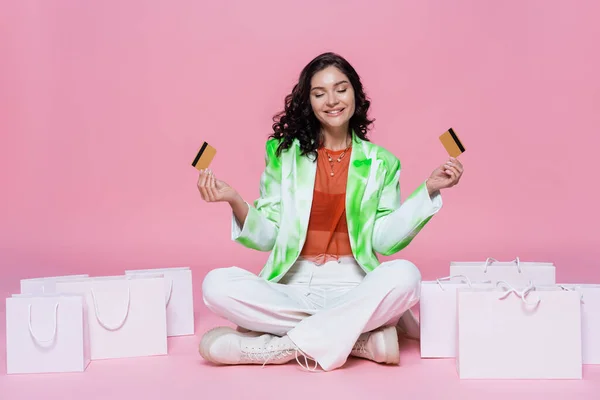 happy woman in blazer holding credit cards while sitting with crossed legs near shopping bags on pink