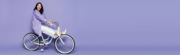full length of amazed young woman in violet raincoat riding bicycle on purple, banner