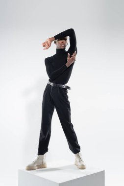 Beyaz kübün üzerinde durup gri renkte poz veren siyahlı Afro-Amerikan bir adam.