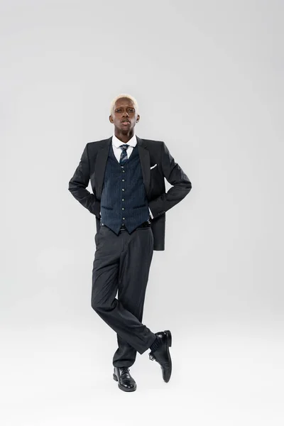 スーツ姿のブロンドアフリカ系アメリカ人男性の完全な長さ灰色で脚を組んで立って — ストック写真