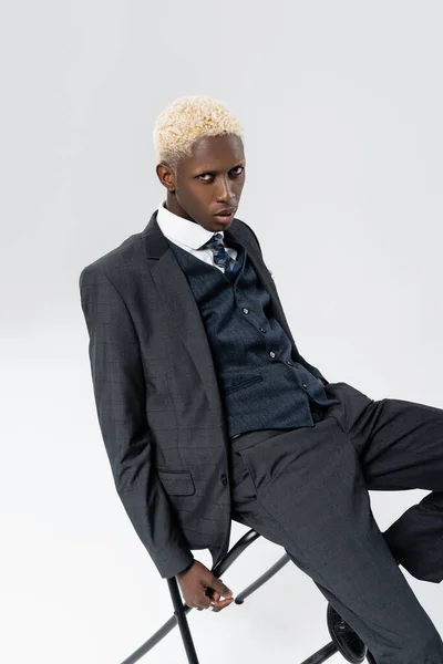 blonde african american man in suit posing on chair isolated on grey