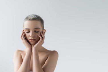 Çıplak omuzları ve kapalı gözleri olan güzel bir kadın Gri renkte izole edilmiş yüzlere dokunuyor.