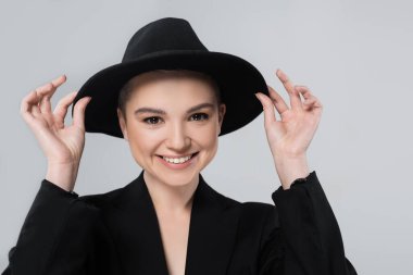 front view of smiling woman adjusting black brim hat isolated on grey clipart