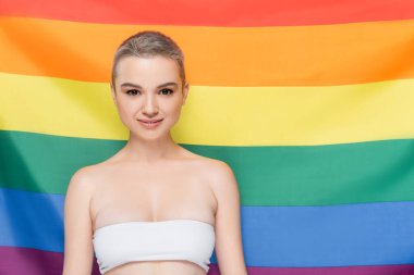 Beyaz elbiseli genç bir kadın arka planda lgbt bayrağı yakınlarında kameraya bakıyor.