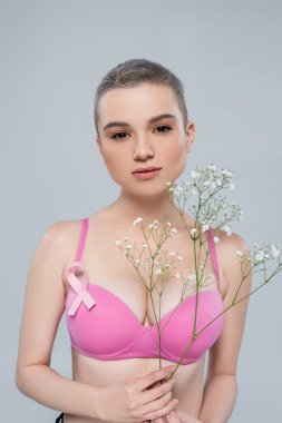 Sütyeninde pembe farkındalık kurdelesi olan genç bir kadın gri üzerinde küçük çigila çiçekleri tutuyor.