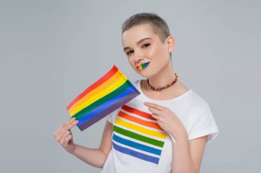 Gökkuşağı renginde dudakları ve küçük lgbt bayrağı olan kadın gri renkli kameraya bakıyor.