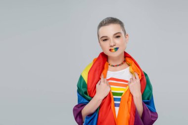 Omuzlarında lgbt bayrağı olan genç ve güzel bir kadın gri kameraya bakıyor.