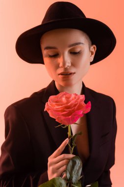 charming woman in black brim hat holding pink rose on coral background clipart
