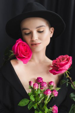 Siyah fötr şapkalı çekici kadın, koyu arkaplanda taze pembe güllerin yanında.