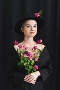 Siyah ceketli ve şapkalı çekici kadın koyu arkaplanda pembe güllerle poz veriyor.