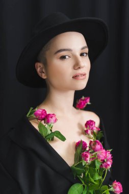 Şapkalı kadın siyah arka planda taze pembe güllerle poz veriyor.