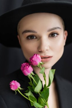 Siyah fötr şapkalı çekici kadın arka planda pembe güllerin yanında kameraya bakıyor.