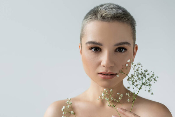 charming woman with naked shoulders looking at camera near small white flowers isolated on grey