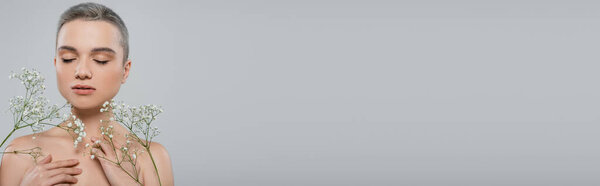 woman with short hair, closed eyes and bare shoulders near branches of white gypsophila isolated on grey, banner