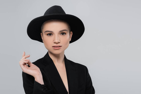 stylish woman in black fedora hat and blazer looking at camera isolated on grey