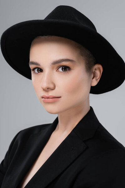 portrait of young woman with natural makeup, wearing black brim hat, looking at camera isolated on grey