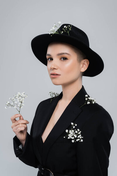 woman with white tiny flowers on black blazer and fedora hat looking at camera isolated on grey