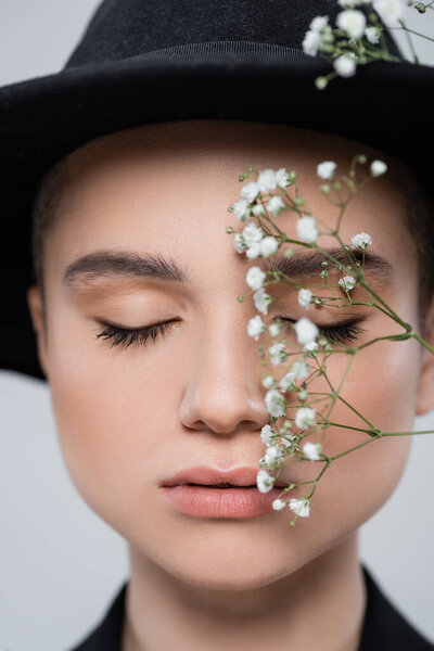 close up portrait of woman with closed eyes and natural makeup near small white flowers isolated on grey