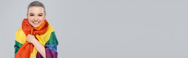 улыбающаяся женщина, обернутая флагом lgbt, глядя на камеру, изолированную на сером, баннере
