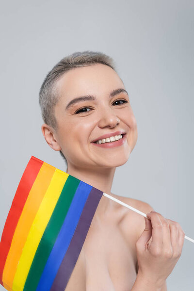 взволнованная женщина смотрит в камеру рядом с маленьким флагом lgbt, изолированным на сером