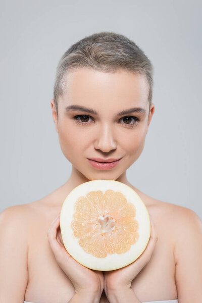 young woman with bare shoulders looking at camera near half of grapefruit isolated on grey