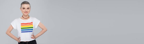 Улыбающаяся женщина в ожерелье и цвета lgbt футболка позирует с руками на бедрах изолированы на сером, баннер