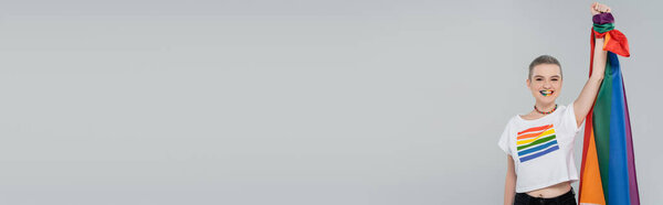 взволнованная женщина с флагом lgbt в поднятой руке, глядя на камеру, изолированную на сером, баннер
