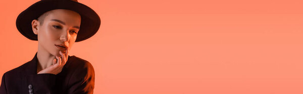 pretty woman in black brim hat touching lips while posing isolated on coral, banner