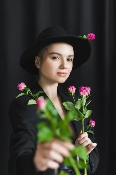 ピンクのバラを背景にピンクのバラを持つつばの帽子と黒のブレザーの可愛い女性 — ストック写真