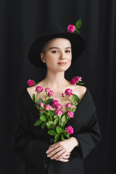魅力的な女性で黒ブレザーとつばの帽子ポージングで新鮮なピンクのバラで暗い背景 — ストック写真