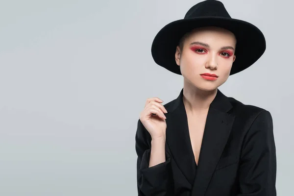 stylish woman in black blazer and fedora hat looking at camera isolated on grey