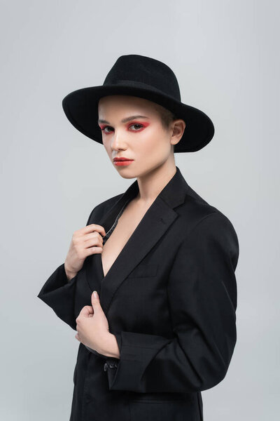 sensual young woman in brim hat touching black blazer while looking at camera isolated on grey