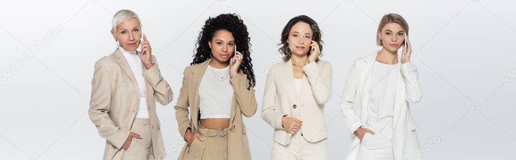 Multiethnic businesswomen in formal wear talking on cellphones isolated on grey, banner 