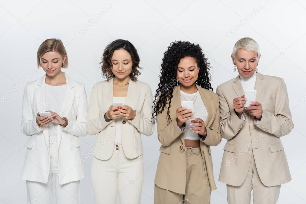 Smiling interracial businesswomen using mobile phones isolated on grey, feminism concept