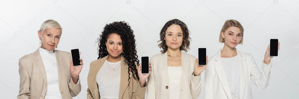 Interracial businesswomen in suits holding smartphones isolated on grey, banner 