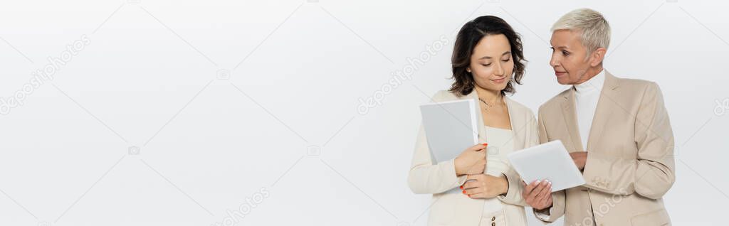 Businesswoman holding digital tablet near colleague with paper folder isolated on grey, banner 