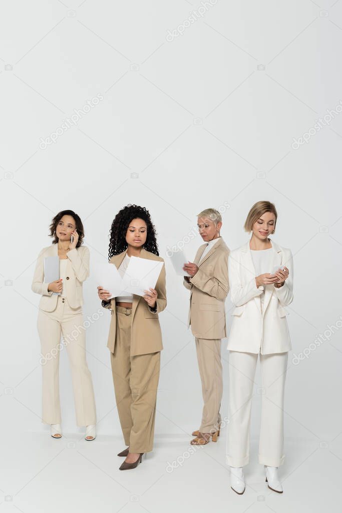 Interracial businesswomen using gadgets and holding papers on grey background 