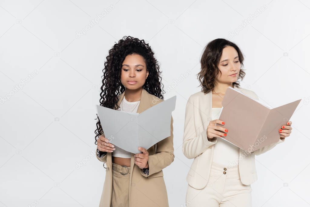 Multiethnic businesswomen in formal wear holding paper folders isolated on grey 