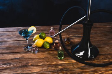 Limonların yanında bir bardak kokteyl ve siyah arka planda ahşap bir yüzey üzerinde dumanlı nargile.