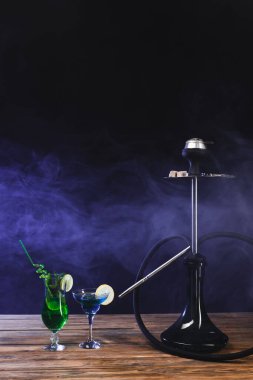 Hookah near glasses of cocktails on wooden surface on black background with smoke clipart