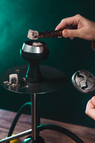 Cropped view of man holding coal in tweezers near hookah on green background with smoke