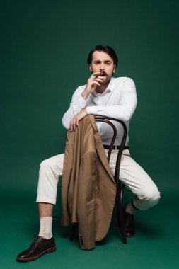 Bıyıklı, puro içen ve koyu yeşil sandalyede oturan bir adam. 