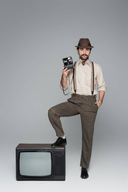 Tam boy erkek retro tarzı kıyafetler ve eski model kamera taşıyan bir şapka ve gri üzerinde antika bir televizyonun yanında poz veriyor.